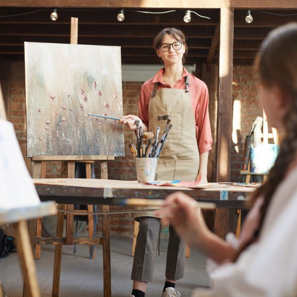 Young teacher standing near the easel pointing at picture and holding art lesson for students at art studio